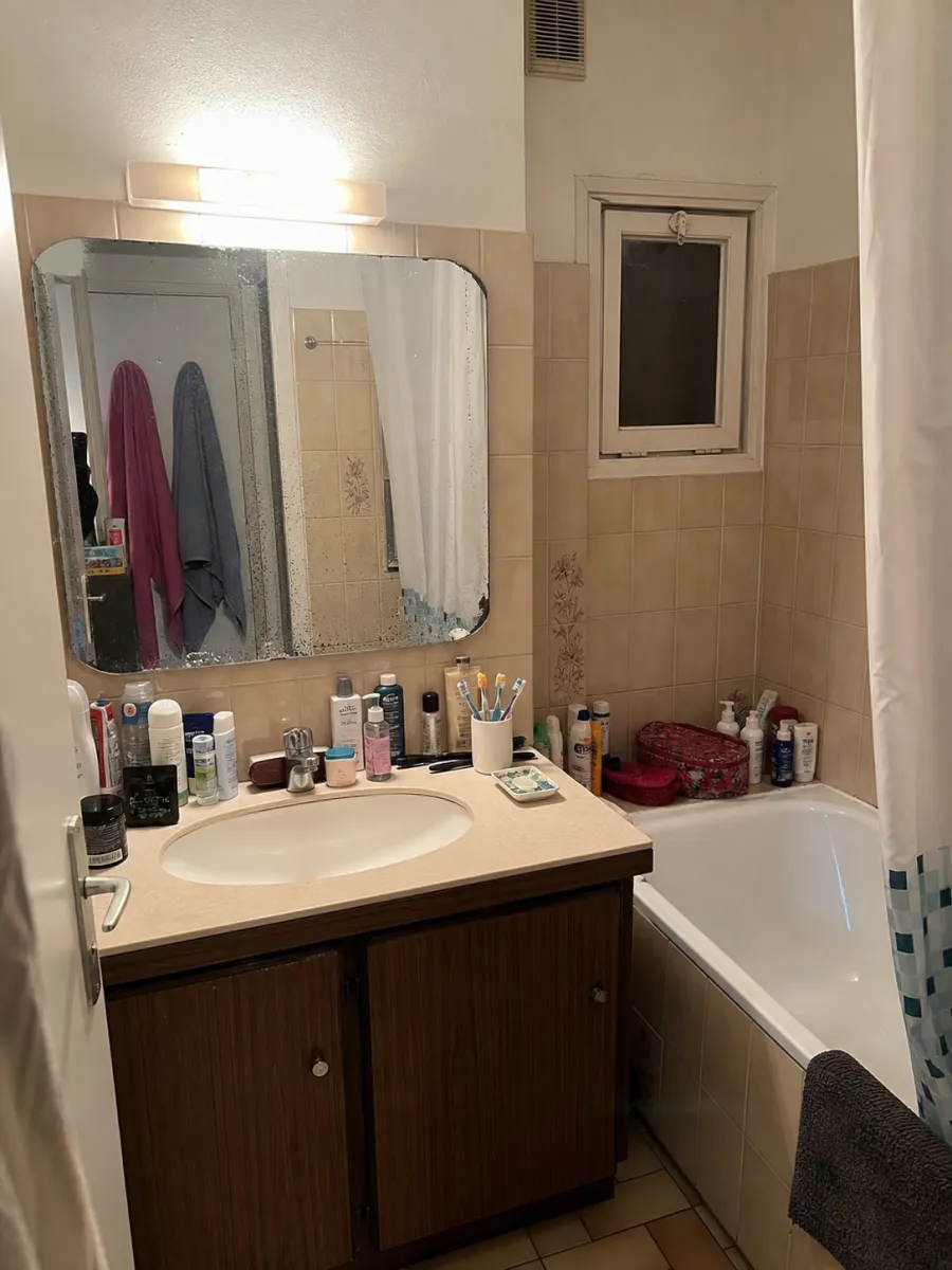 Salle de bains datée avant home staging : faïence beige, meuble vasque sombre, rebord du lavabo encombré de flacons